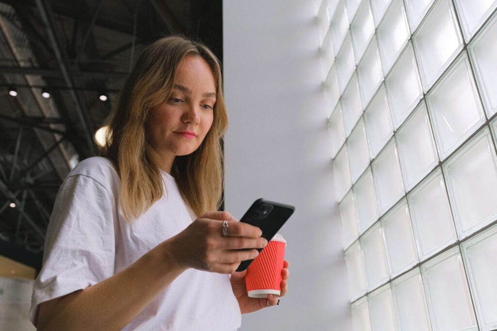 Žemo kampo vaizdas, kuriame jauna šviesiaplaukė moteris, dėvinti baltus marškinėlius, žiūri į savo juodą išmanųjį telefoną ir rankoje laiko raudoną išsineštinės kavos puodelį. Ji stovi šalia didelės stiklo blokelių sienos.