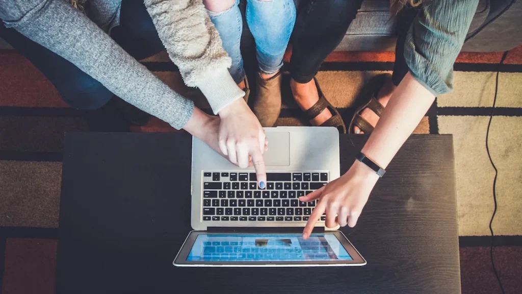 Trys žmonės susibūrę prie nešiojamojo kompiuterio rodo į ekraną, kartu naršydami internete.